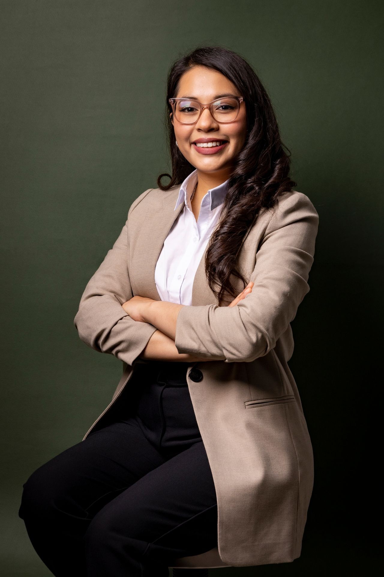 Person in a beige blazer and white shirt sitting with arms crossed against a dark green background.