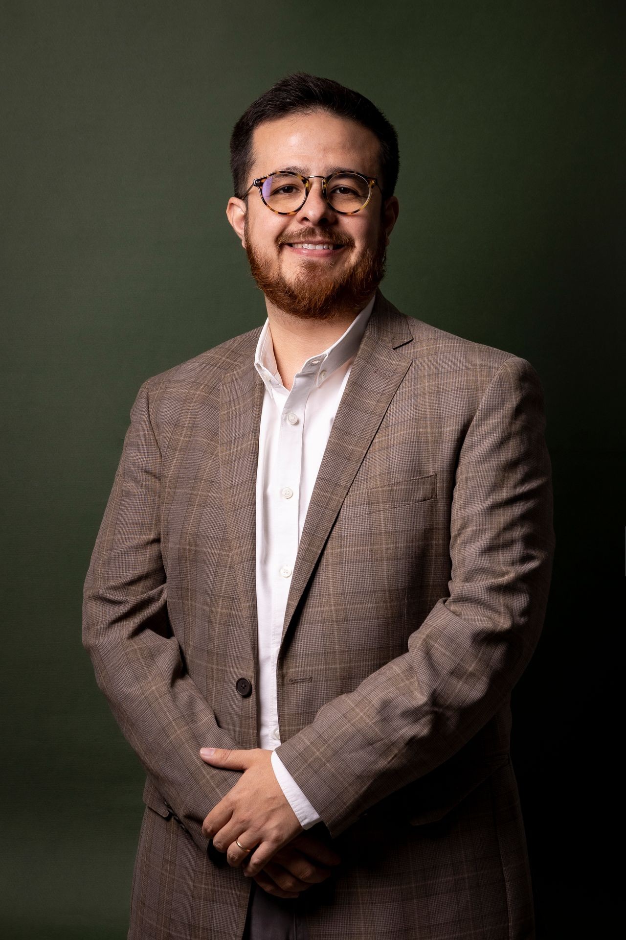 Person wearing a brown plaid suit with a white dress shirt, standing against a dark green background.