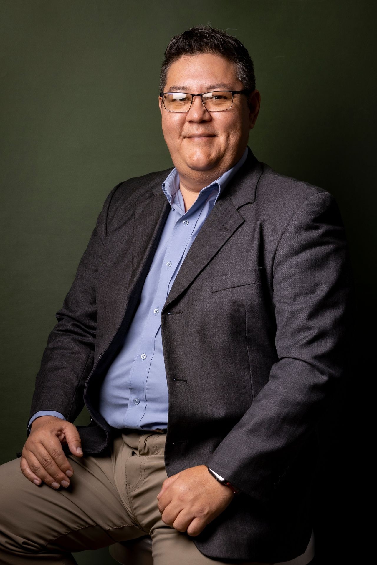 Person posing in a dark suit and blue shirt against a plain dark background.