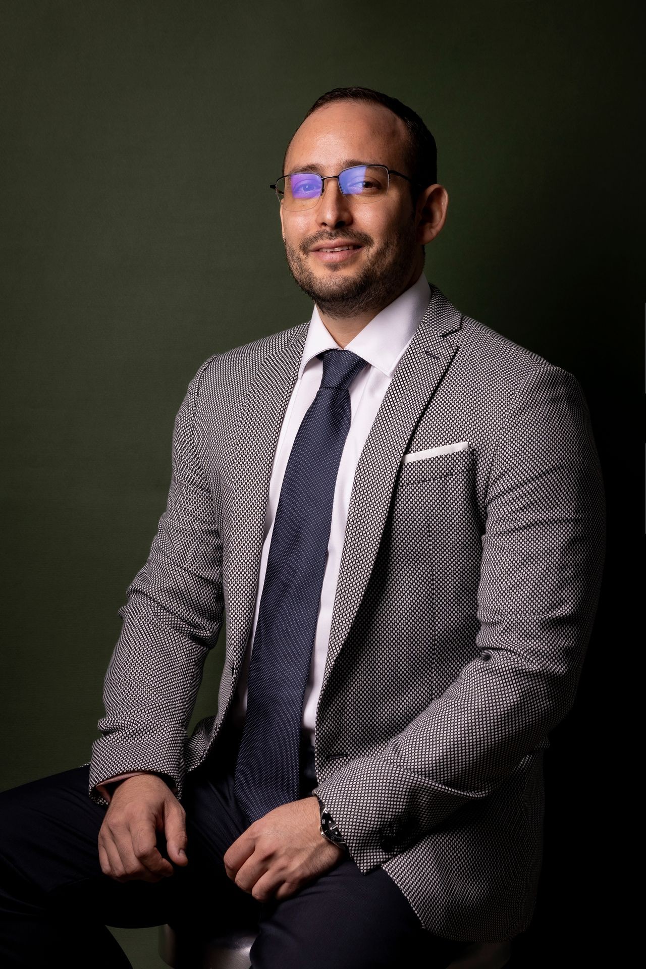 Man in a houndstooth blazer and glasses, seated against a dark green background.