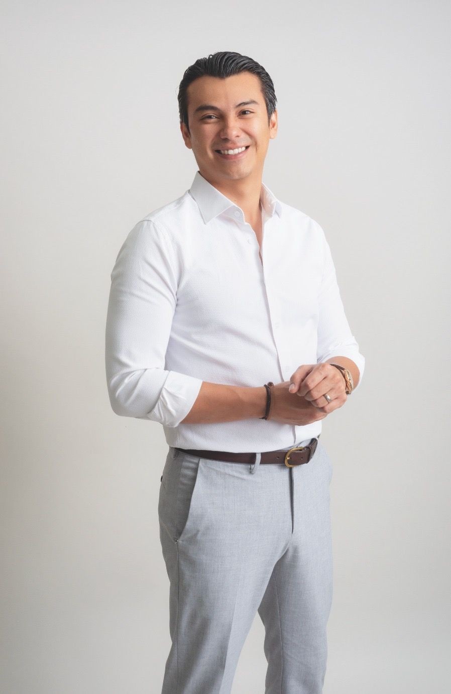 Smiling man in white shirt and gray pants posing with clasped hands in a studio setting.