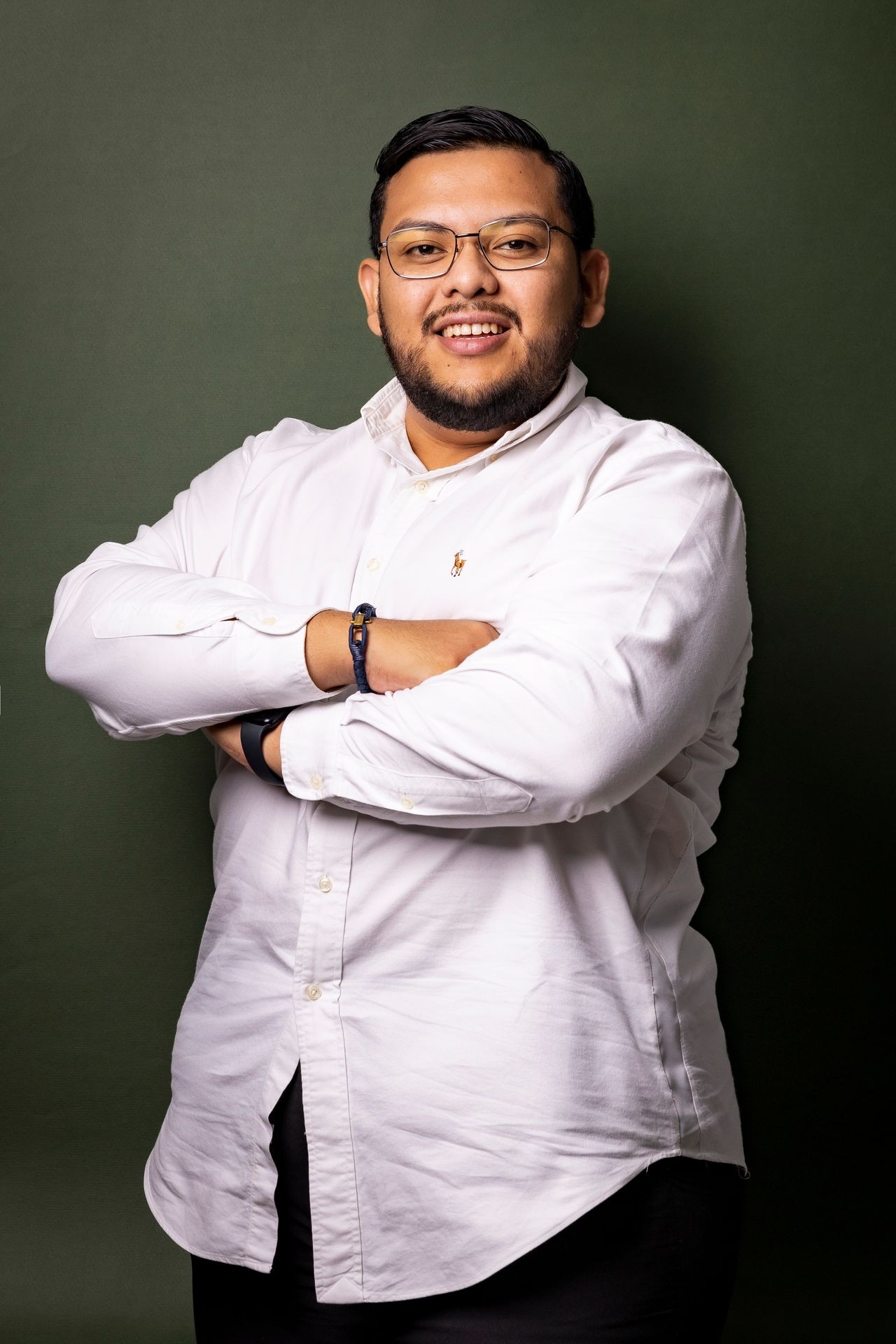 Man in white button-up shirt with crossed arms and glasses smiling against a dark green background.