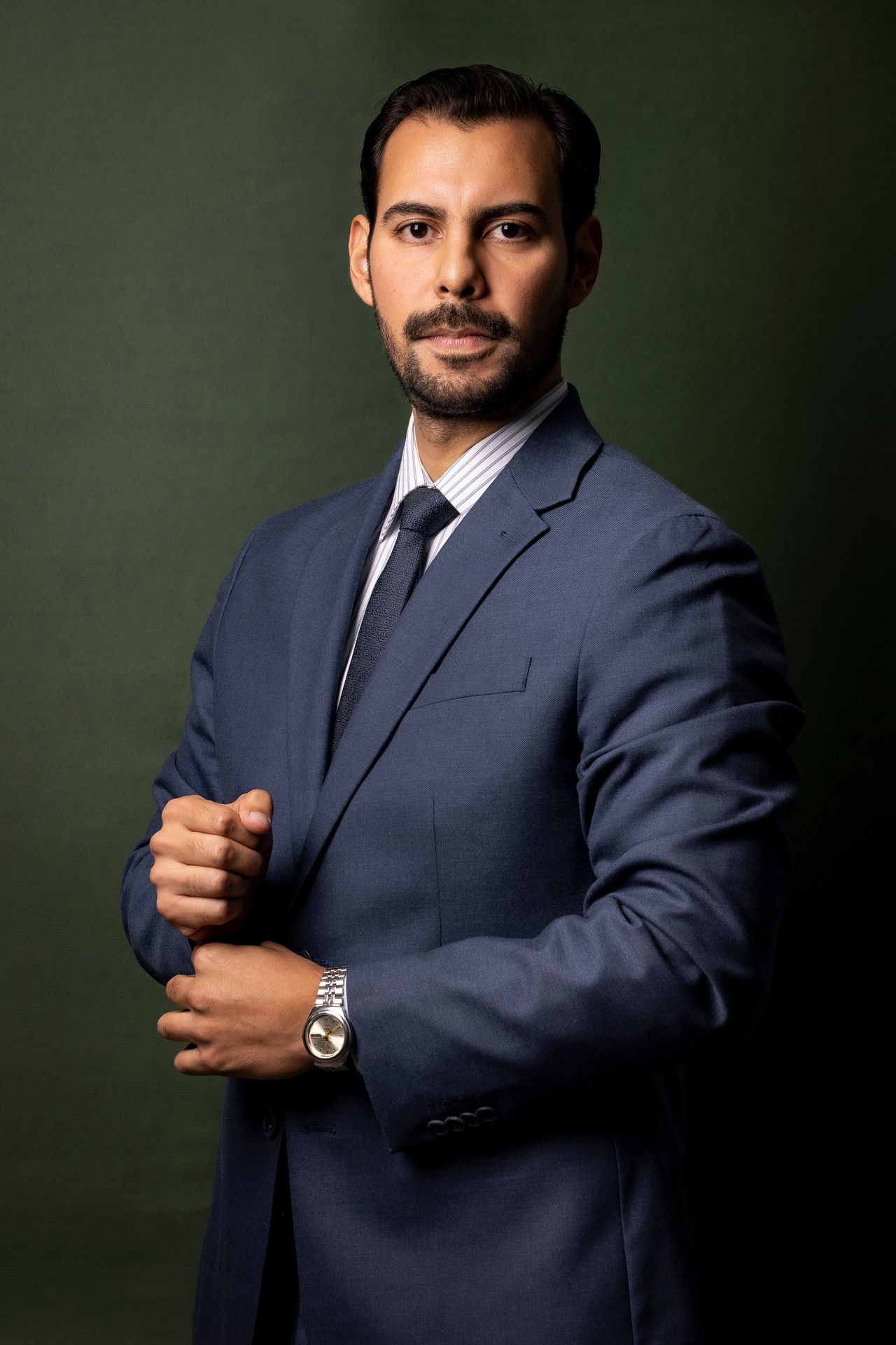 Man in a blue suit posing confidently against a dark green background.