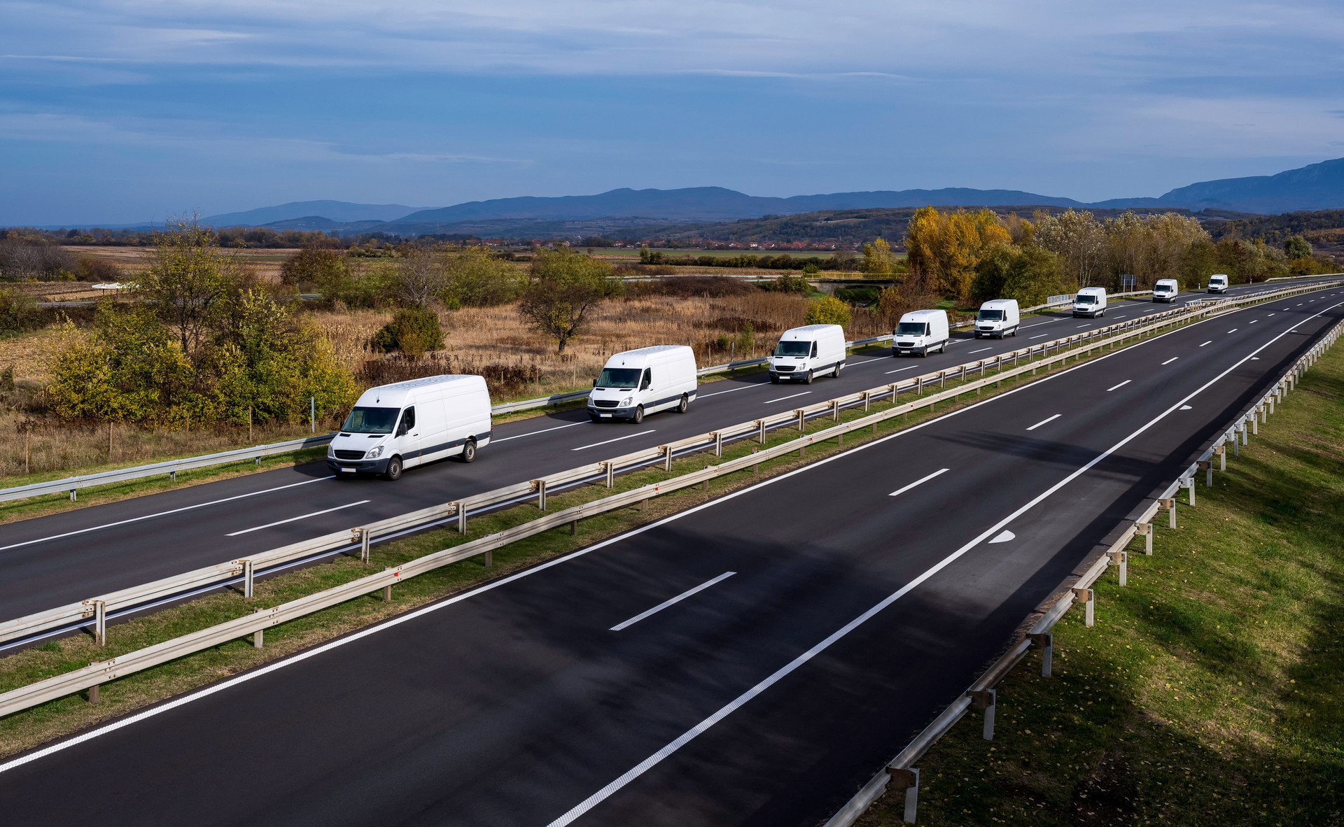 Ocho minivans blancas se mueven en un convoy en la carretera. Van de reparto blanca en la carretera. Blanco moderno entrega pequeña carga carga furgoneta de mensajería que se mueve rápido en la carretera de la autopista al suburbio urbano de la ciudad.