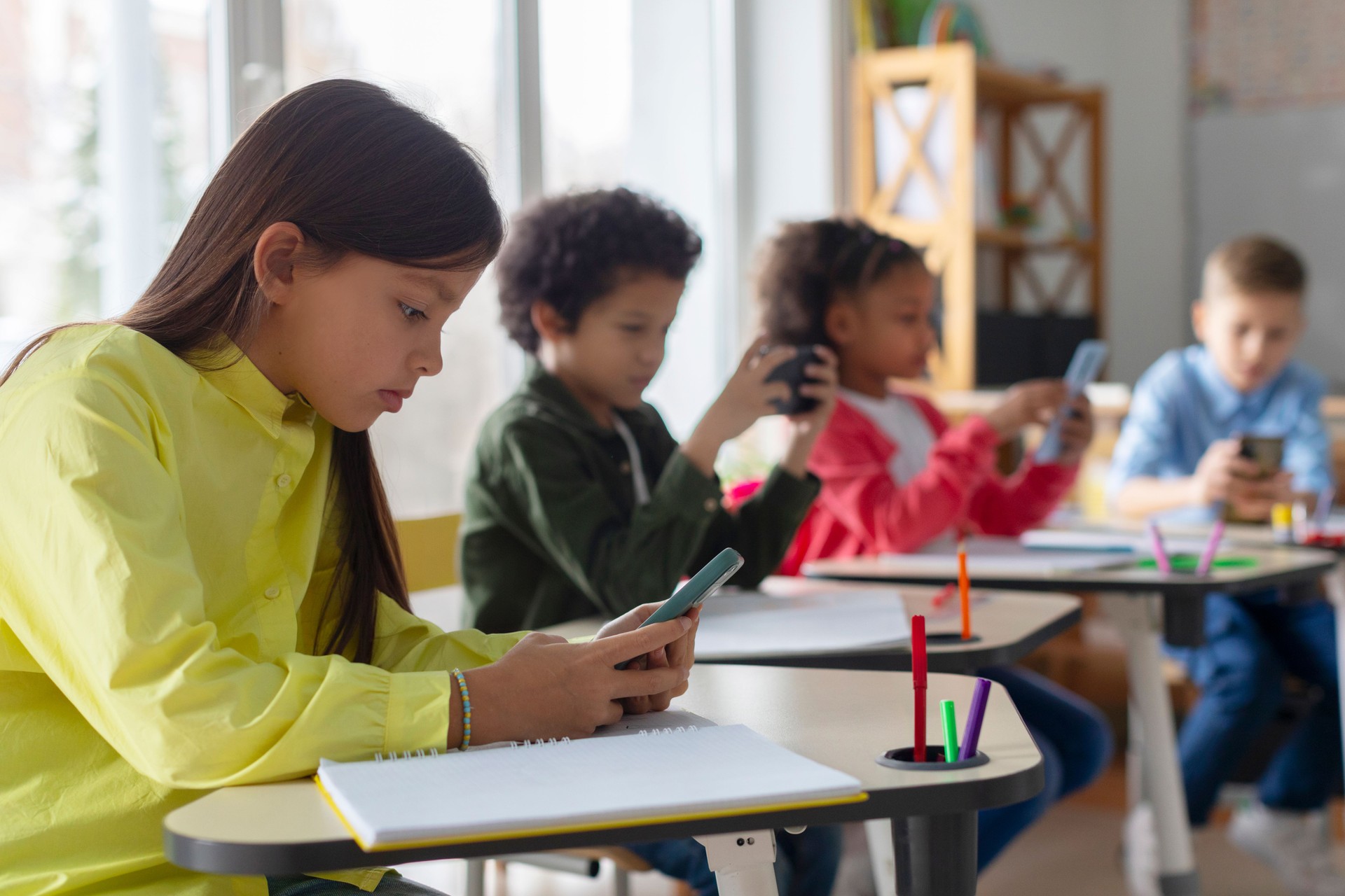 Niños multiétnicos diversos, estudiantes que tienen un descanso y usan teléfonos inteligentes, se sientan en los escritorios en el aula, niños juegan y revisan las redes sociales en la escuela.